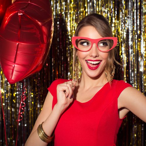 Young woman wearing elegant red dress celebrating or having party in front of fringe curtain. She is holding photo booth fake glasses and posing at camera.